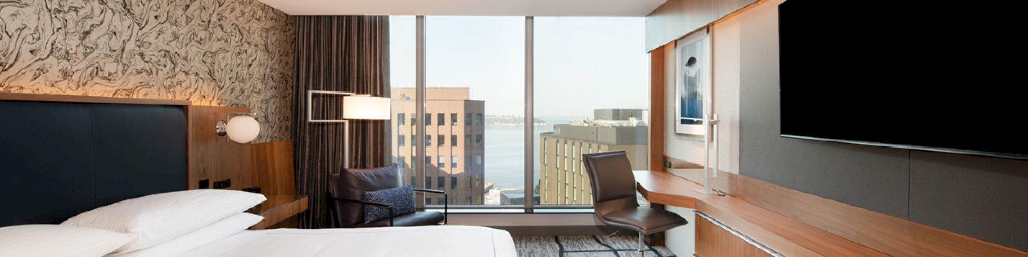 A modern hotel room with a large bed, flat-screen TV, desk, and a window view of city buildings and water.