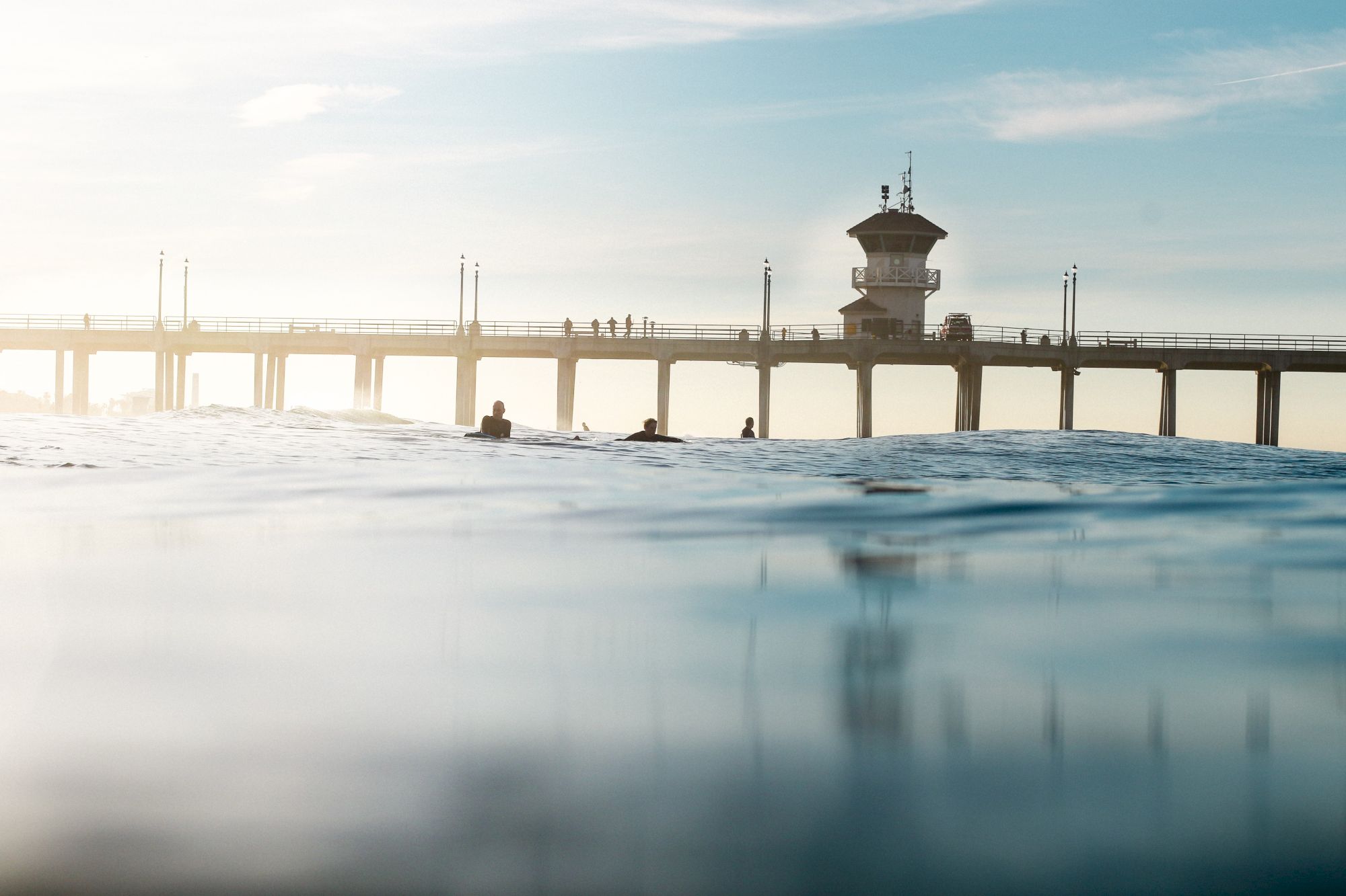 A pier extends over the ocean with people walking on it, while surfers ride waves in the foreground under a clear sky.